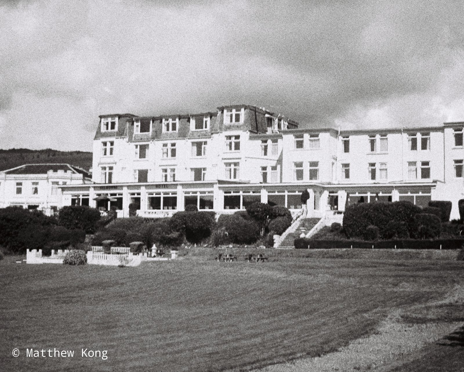 A long white hotel behind clipped hedges and a wide empty lawn, under a heavy sky.