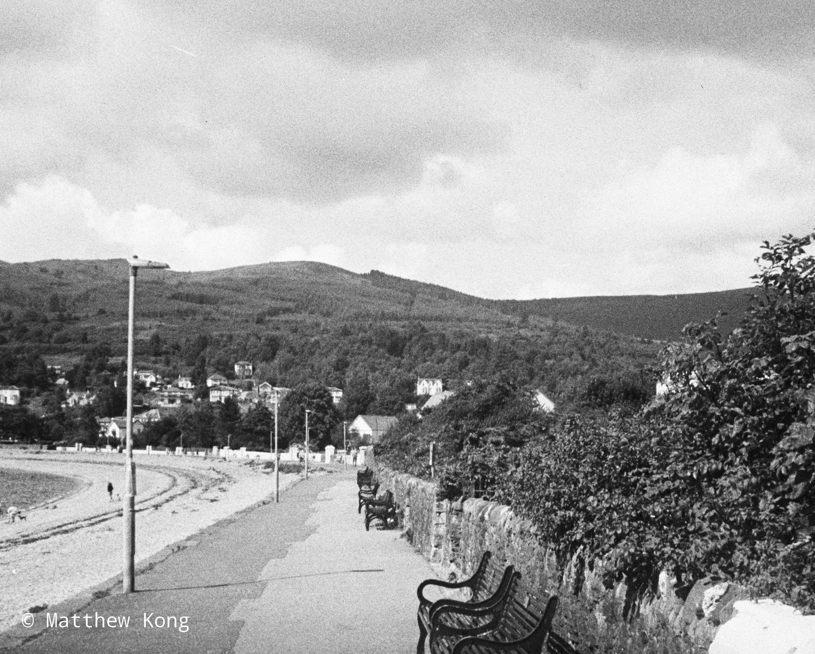 An empty promenade curving along the shore, benches against the sea wall and wooded hills beyond.