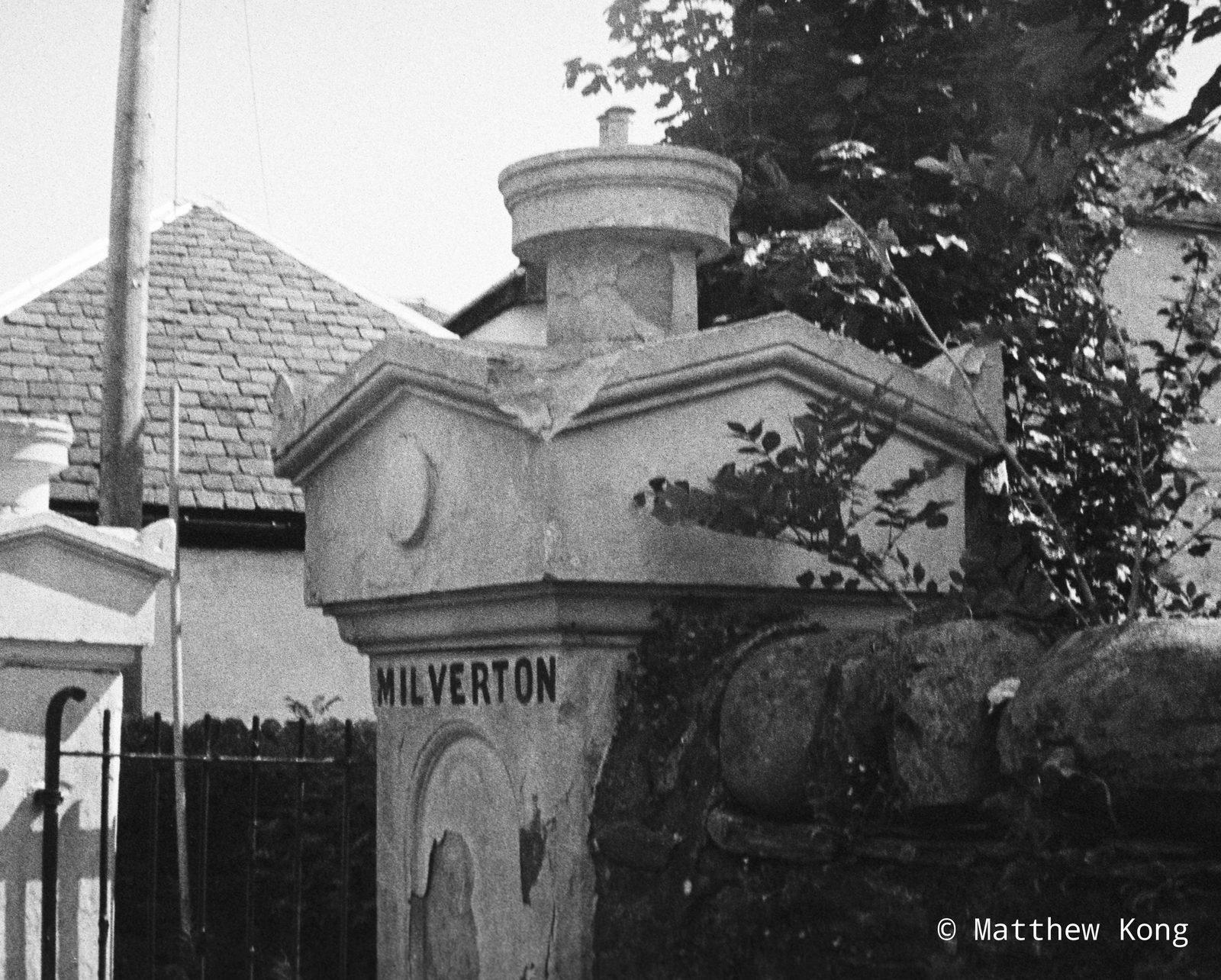 A stone gatepost with MILVERTON cut into it, ivy spilling over the wall beside it and a slate-roofed lodge behind.