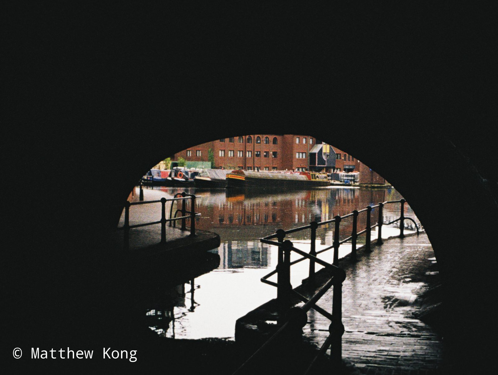 Moored narrowboats and a red brick warehouse, framed by the dark arch of a canal bridge.