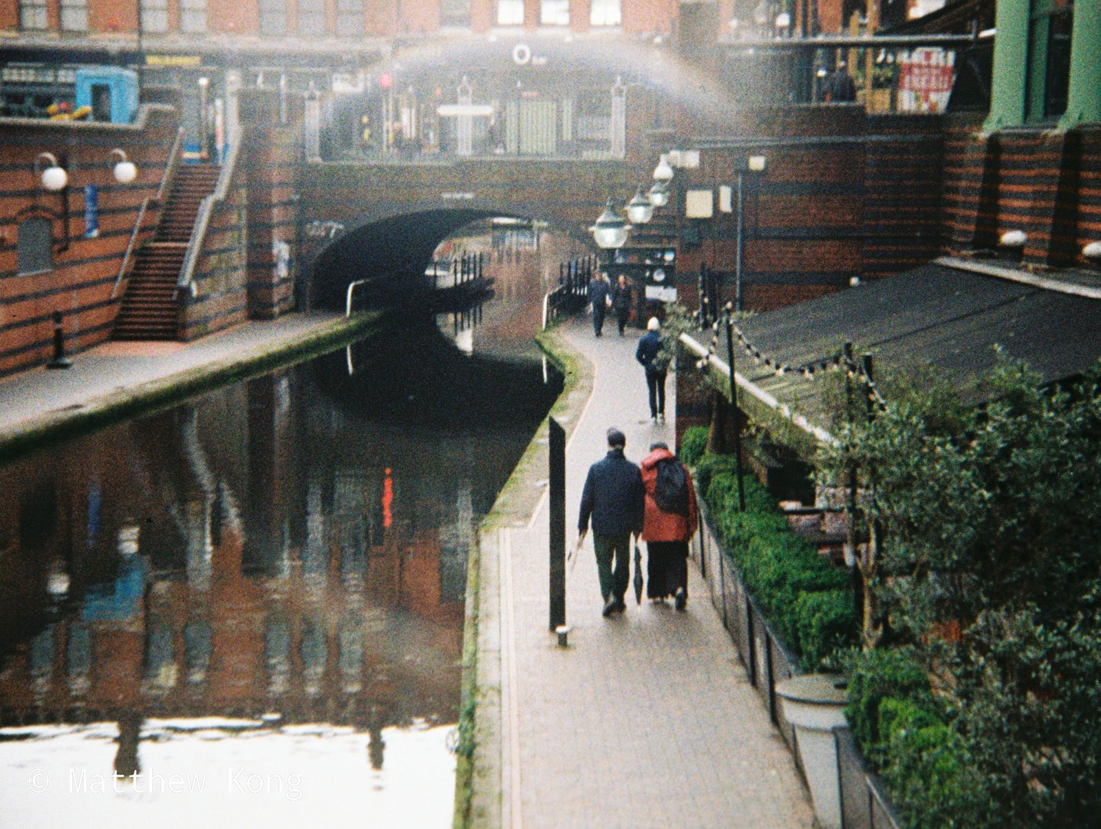 Two people walking away along the towpath towards a bridge, reflected in the water.